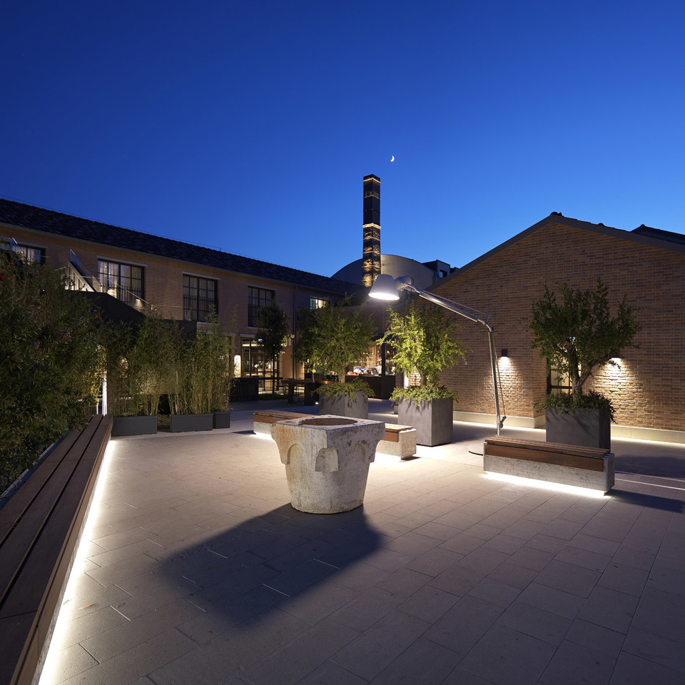 Illuminated outdoor courtyard at dusk with LED strip lighting along benches, uplighting on trees, and a large articulated floor lamp