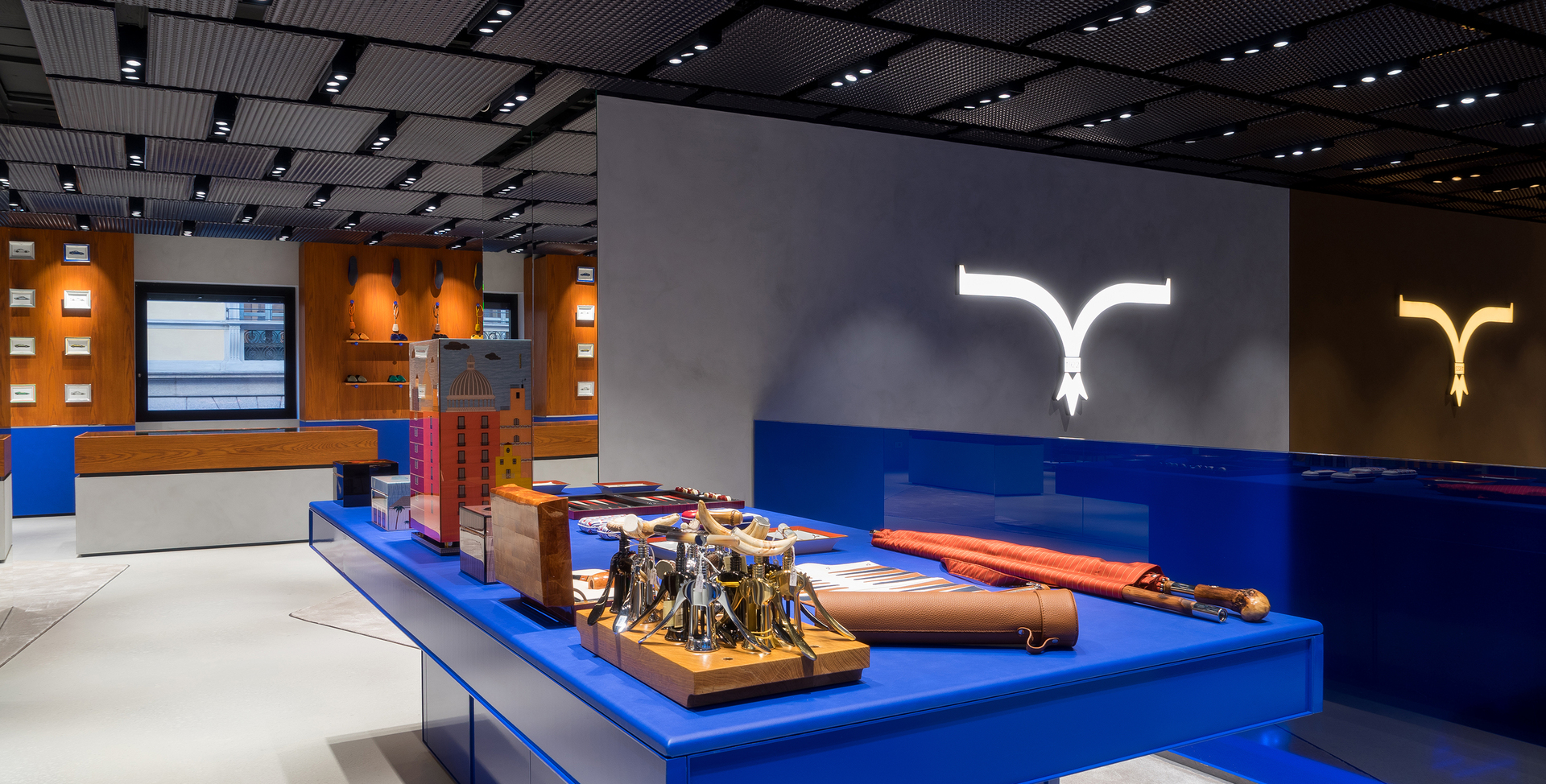 Modern retail interior with blue display table, orange wood panels, recessed ceiling spotlights, and illuminated white logo signs on gray wall.