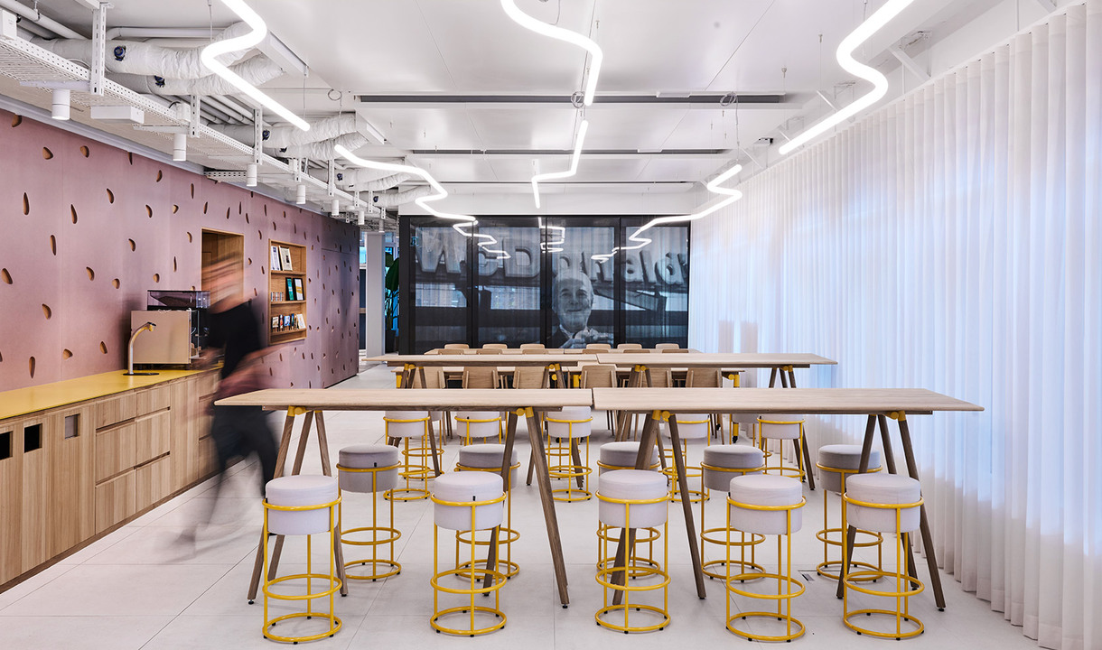 Spazio cucina aziendale con tavoli alti, sgabelli gialli e luci LED curve a soffitto.