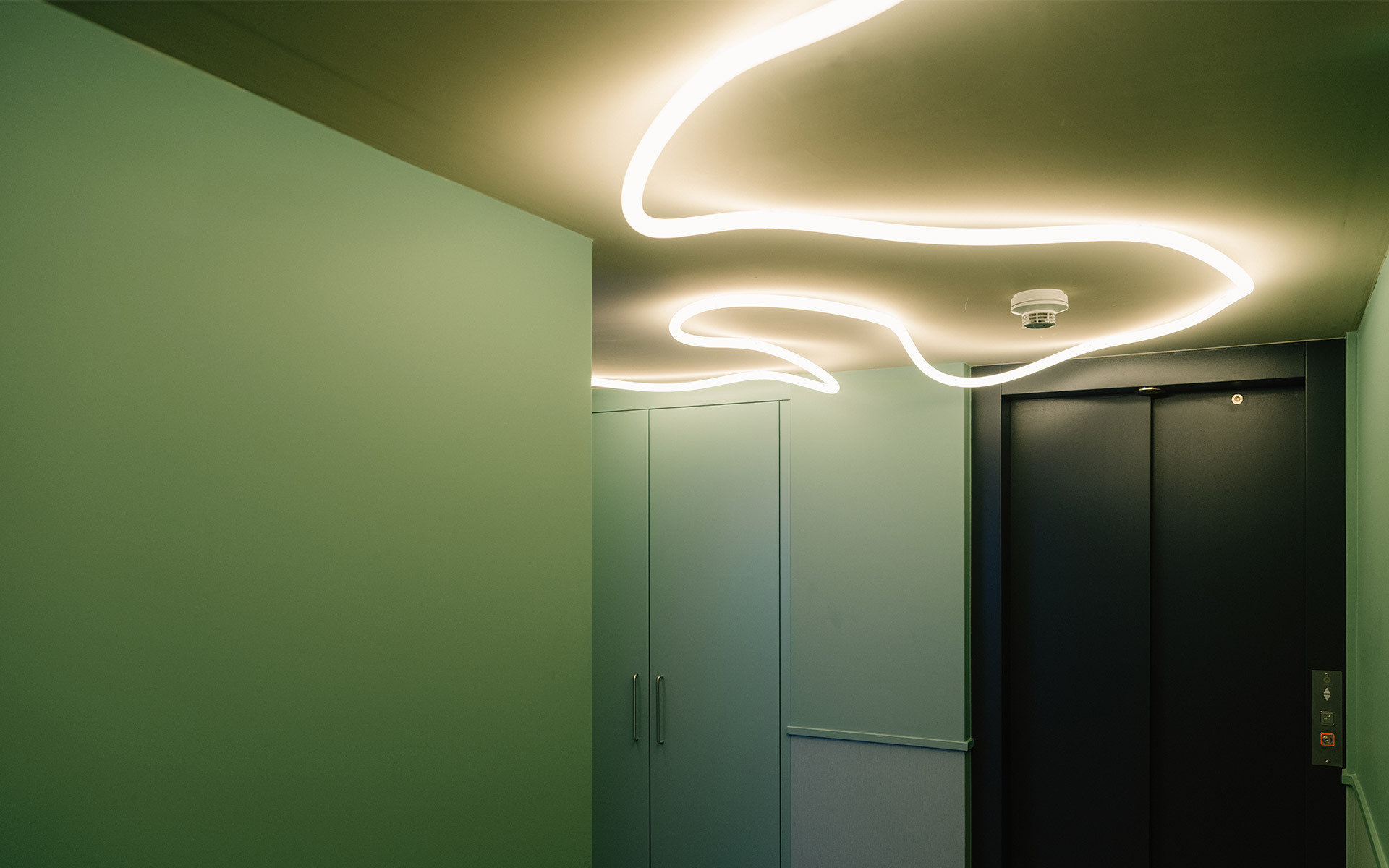 Strisce LED curve che formano onde organiche su un soffitto verde, illuminando un corridoio con porte dell'ascensore.