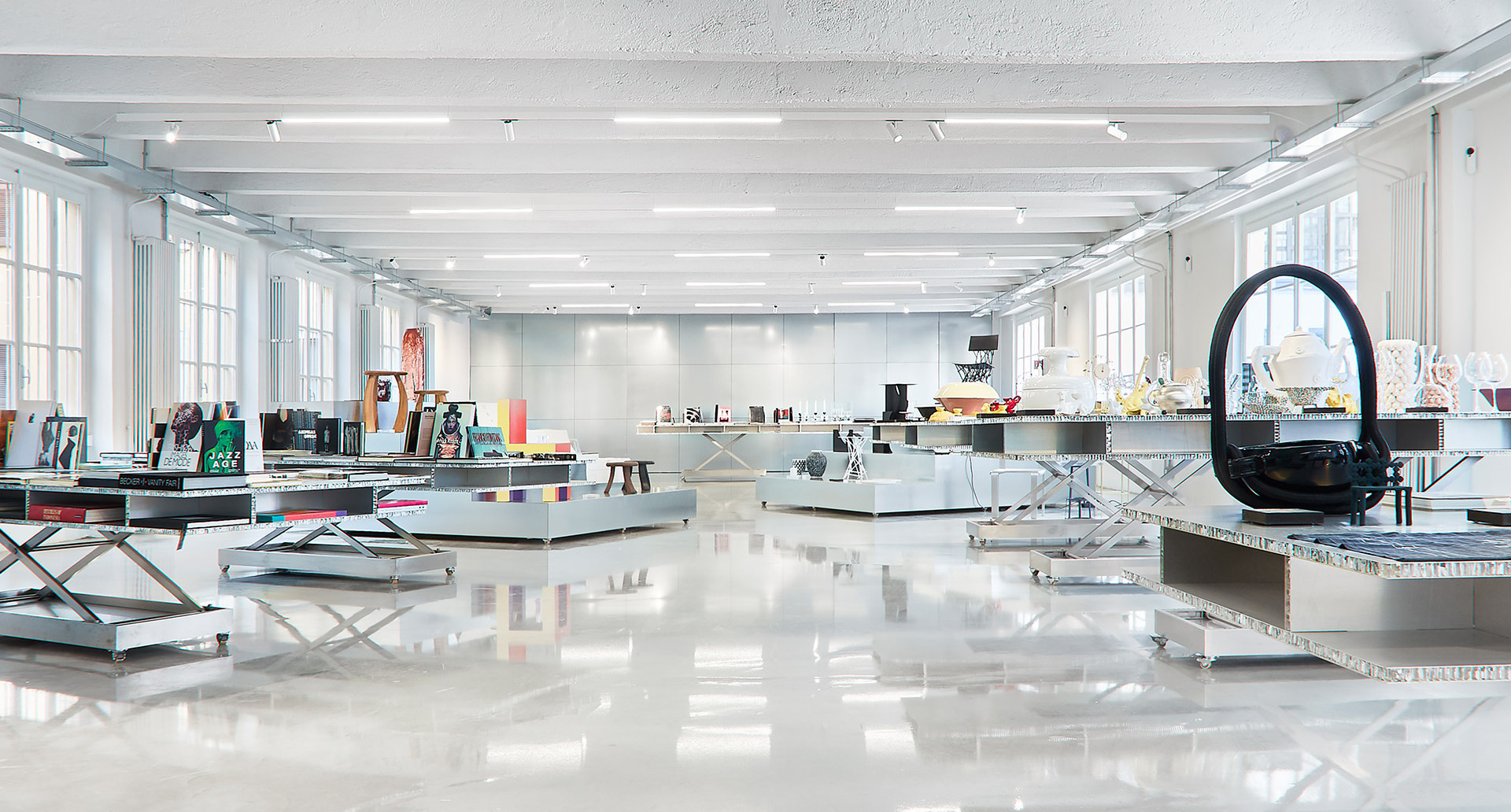 Showroom aperto e luminoso con soffitti bianchi, illuminazione a incasso, pavimenti lucidi e tavoli espositivi con oggetti di design e lampade.