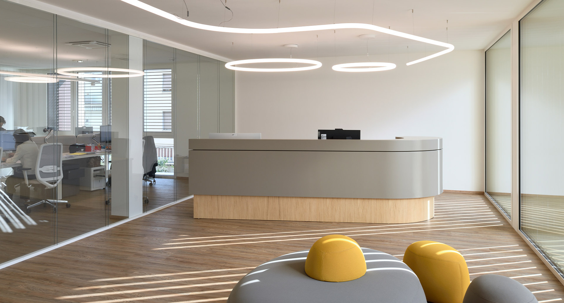 Area reception della nuova sede direzionale Fornaroli Polymers con banco curvo, pavimento in legno e pareti vetrate, illuminata dal sistema a sospensione Alphabet of Light.