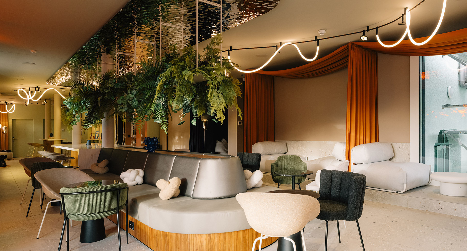 Ampio lounge con sedute imbottite, piante sospese, tende arancioni e sistema Sylt a soffitto con linee luminose.