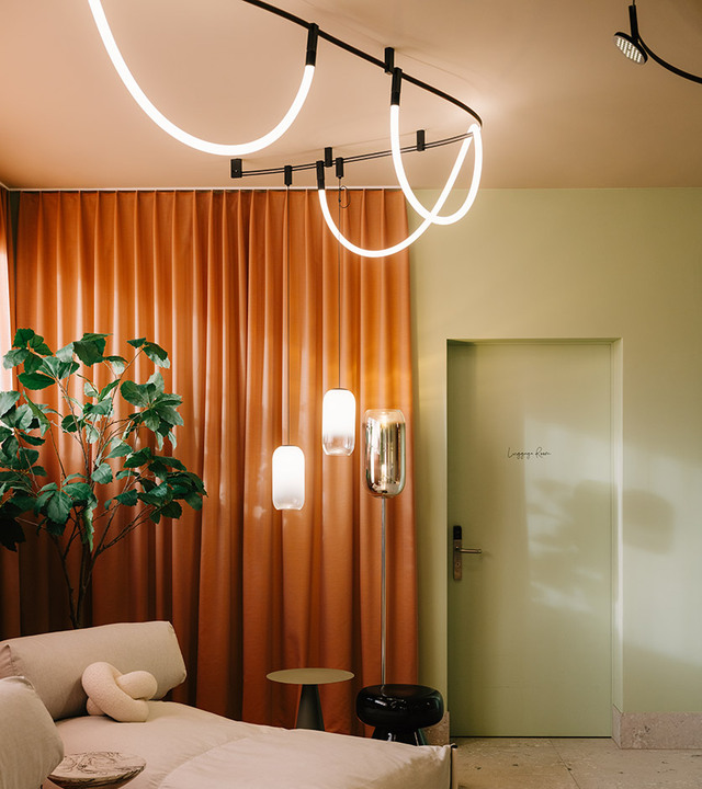 Lounge con tende arancioni, pareti verde chiaro, divano chiaro, lampade Gople Floor sullo sfondo e sistema Sylt a soffitto.