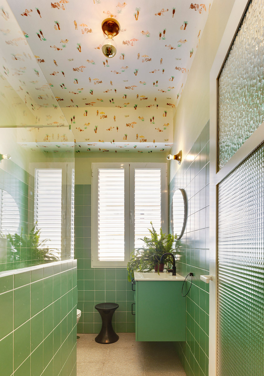 Bagno stretto con rivestimento verde, mobile lavabo sospeso, piante e soffitto decorato a fantasia.