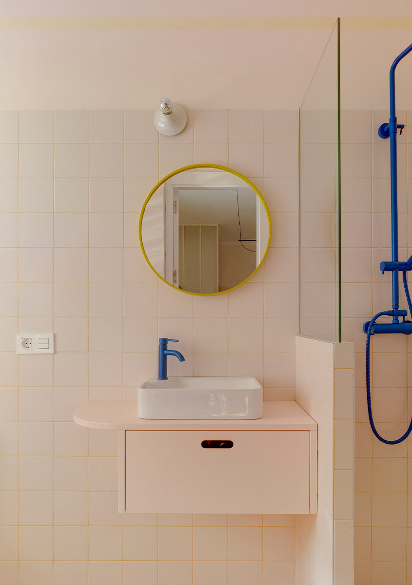 Dettaglio del bagno con lavabo sospeso, specchio rotondo con cornice gialla e rubinetteria blu.