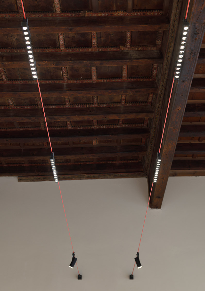 Moduli luminosi lineari e faretti orientabili di Funivia sospesi sotto un soffitto storico decorato in legno.