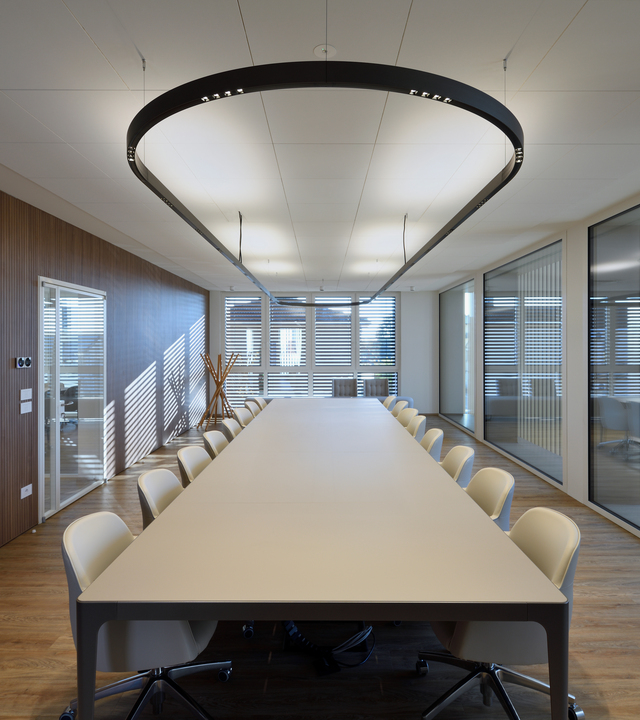 Sala riunioni moderna con pareti vetrate, grande tavolo centrale e sedie chiare, illuminata dal sistema luminoso A.24 a sospensione.