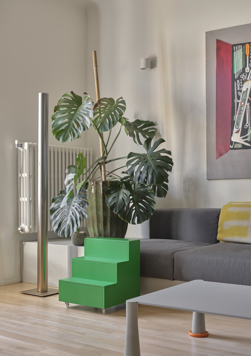 Angolo del soggiorno con lampada da terra Ilio 10 di Artemide, grande monstera in vaso e tavolino verde a gradini accanto al divano.