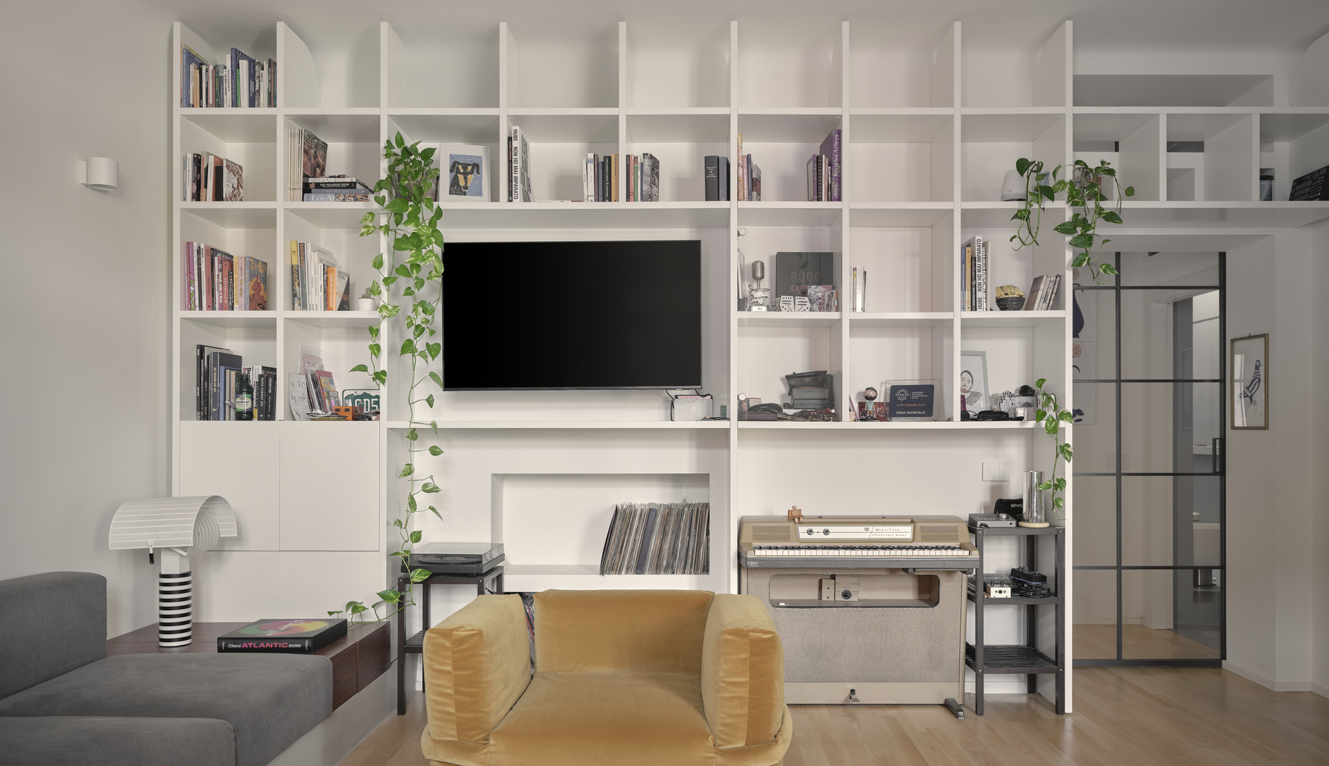 Vista frontale della zona living con libreria a parete, poltrona in velluto ocra e lampada Shogun di Artemide.