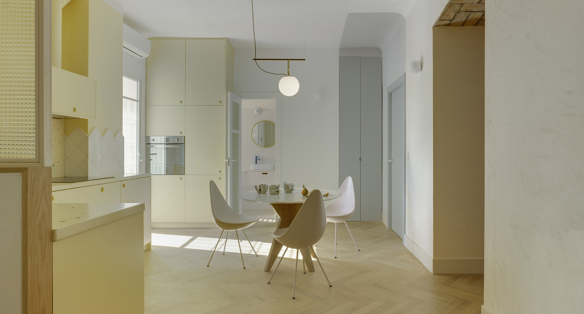 Vista della cucina con zona pranzo, pavimento in legno a spina di pesce e arredi in tonalità crema, il tutto illuminato da una lampada a sospensione.
