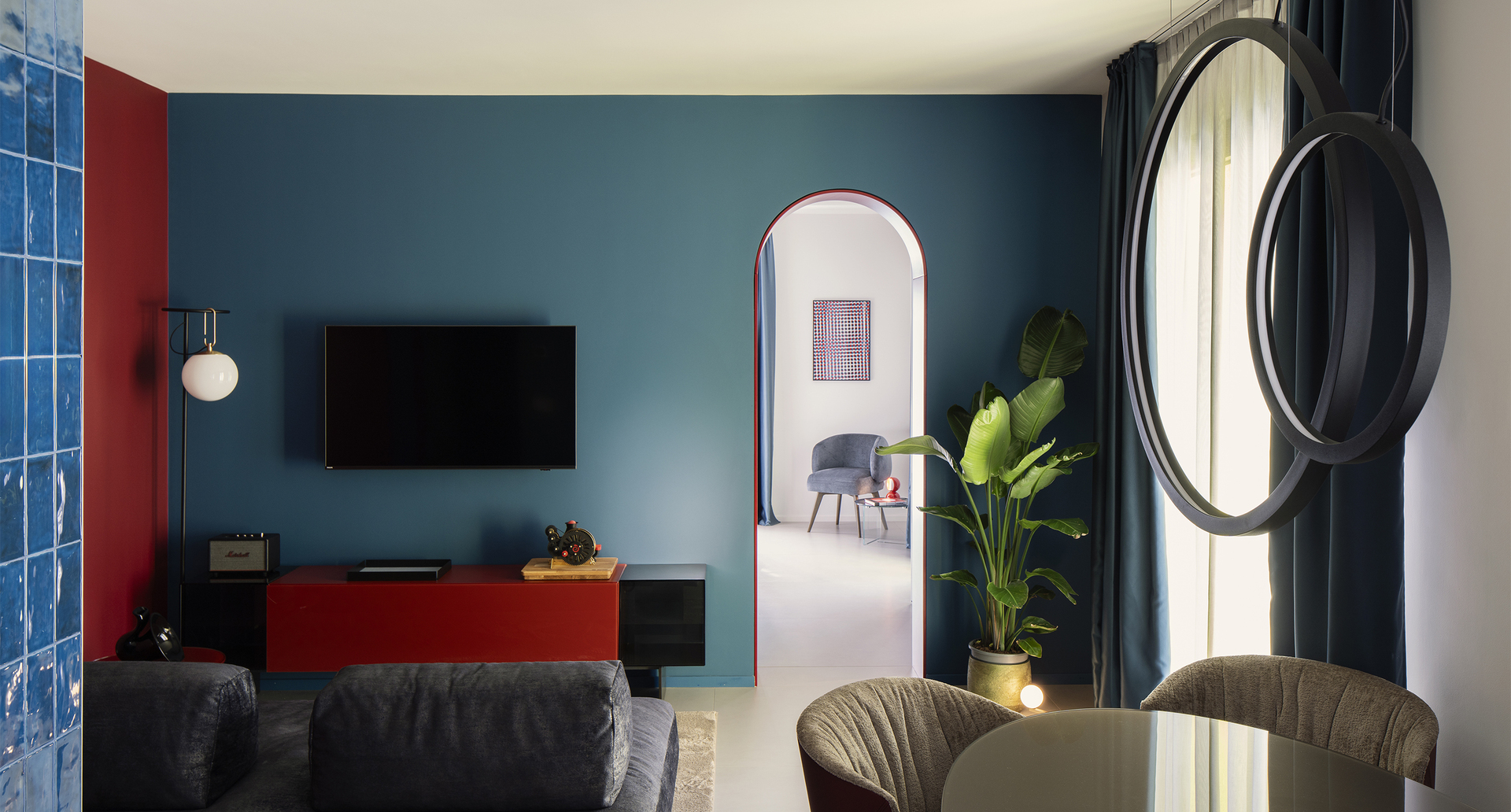 Interno della suite: il salotto accogliente con tavolo illuminato dalle sospensioni “O”, divano elegante e lampada da terra nh vicino al mobile TV.