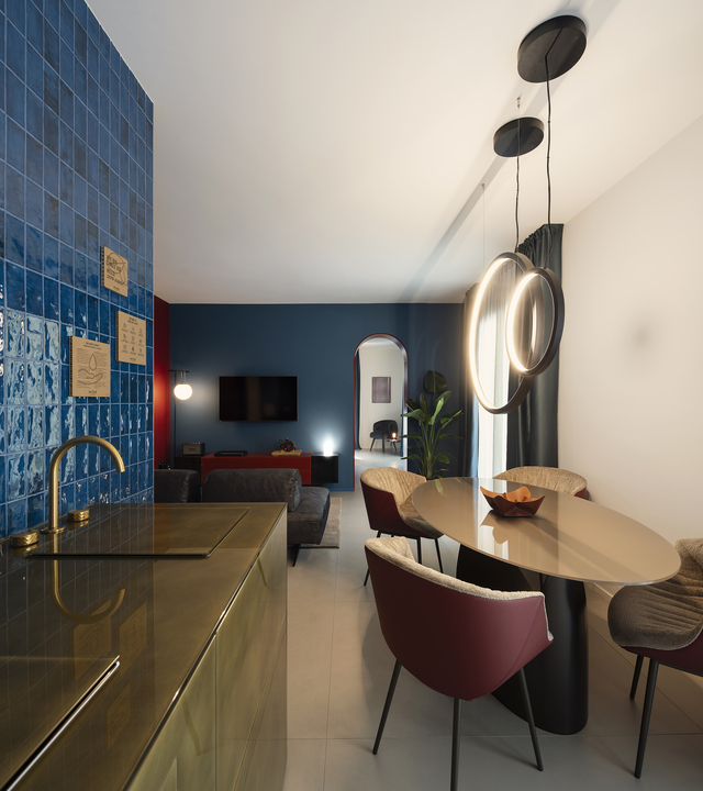 Vista del salotto con il tavolo della zona giorno illuminato dalle sospensioni “O”; sullo sfondo si distinguono il divano e la TV, mentre in primo piano appare la cucina.