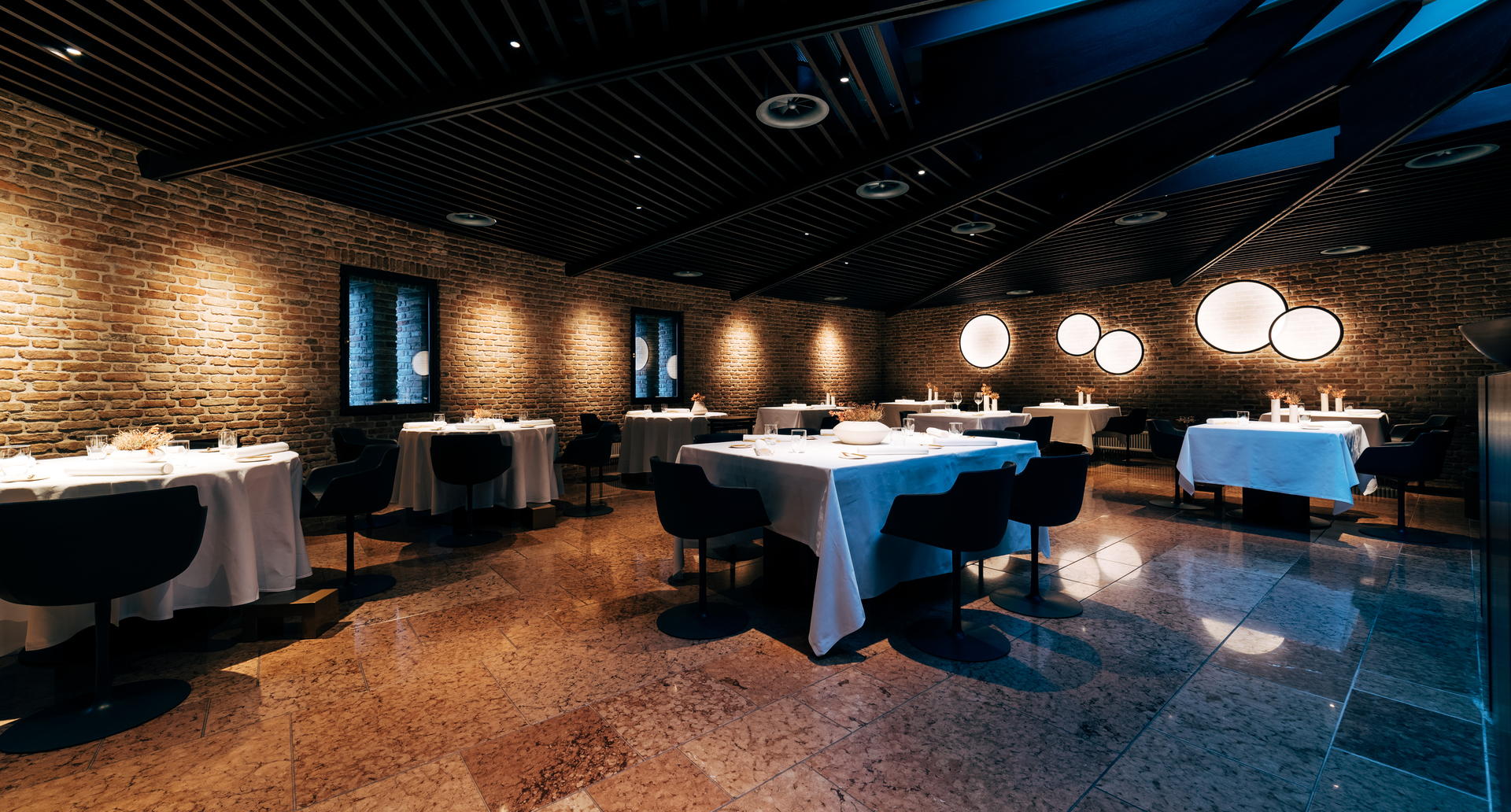 Elegante sala da pranzo con pareti in mattoni a vista, pavimento in marmo, tavoli con tovaglie bianche e luci da parete circolari Artemide che creano un'atmosfera calda.