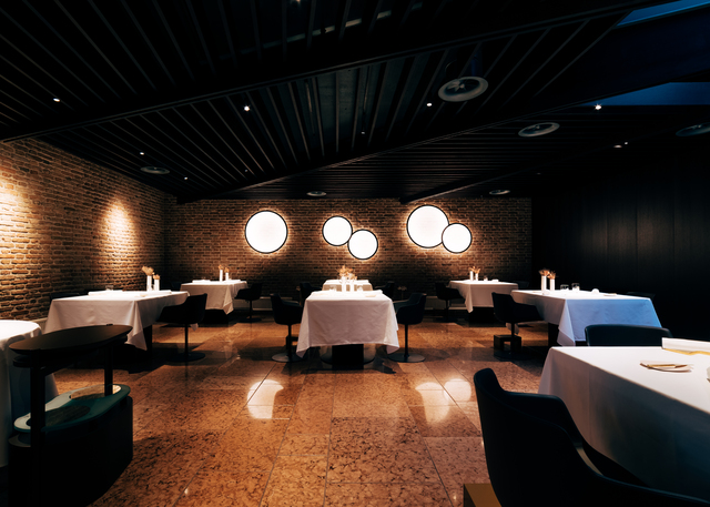 Sala da pranzo elegante con soffitto scuro, pareti in mattoni, tavoli con tovaglie bianche, candele e luminarie circolari a parete che diffondono luce calda.