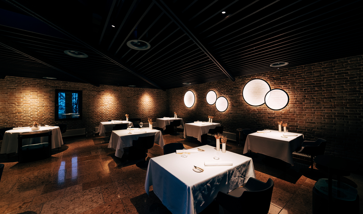 Sala da pranzo elegante con pareti in mattoni, soffitto scuro, tavoli con tovaglie bianche e luci da parete circolari Artemide che creano un'illuminazione calda.