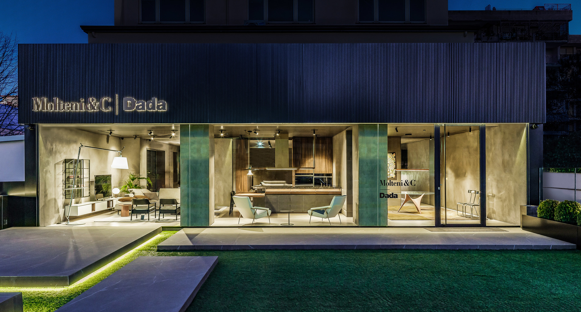 Molteni&C and Dada showroom exterior at dusk, illuminated interiors showcasing furniture through large glass windows with warm lighting