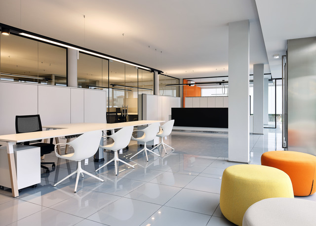 Ufficio open space moderno con luci lineari a soffitto Artemide, postazioni bianche, pareti in vetro e sedute lounge colorate in giallo e arancione