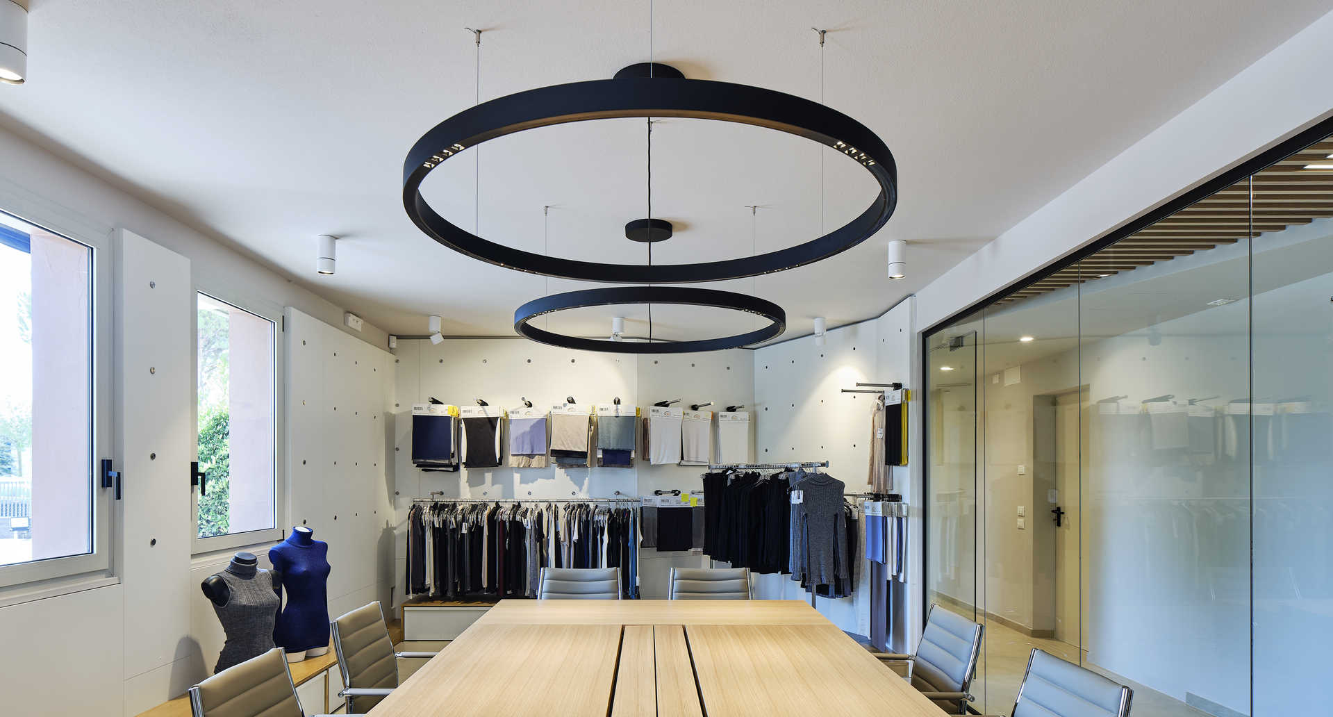Two black circular ring pendant lights suspended over a wooden conference table in a clothing showroom with garment racks.
