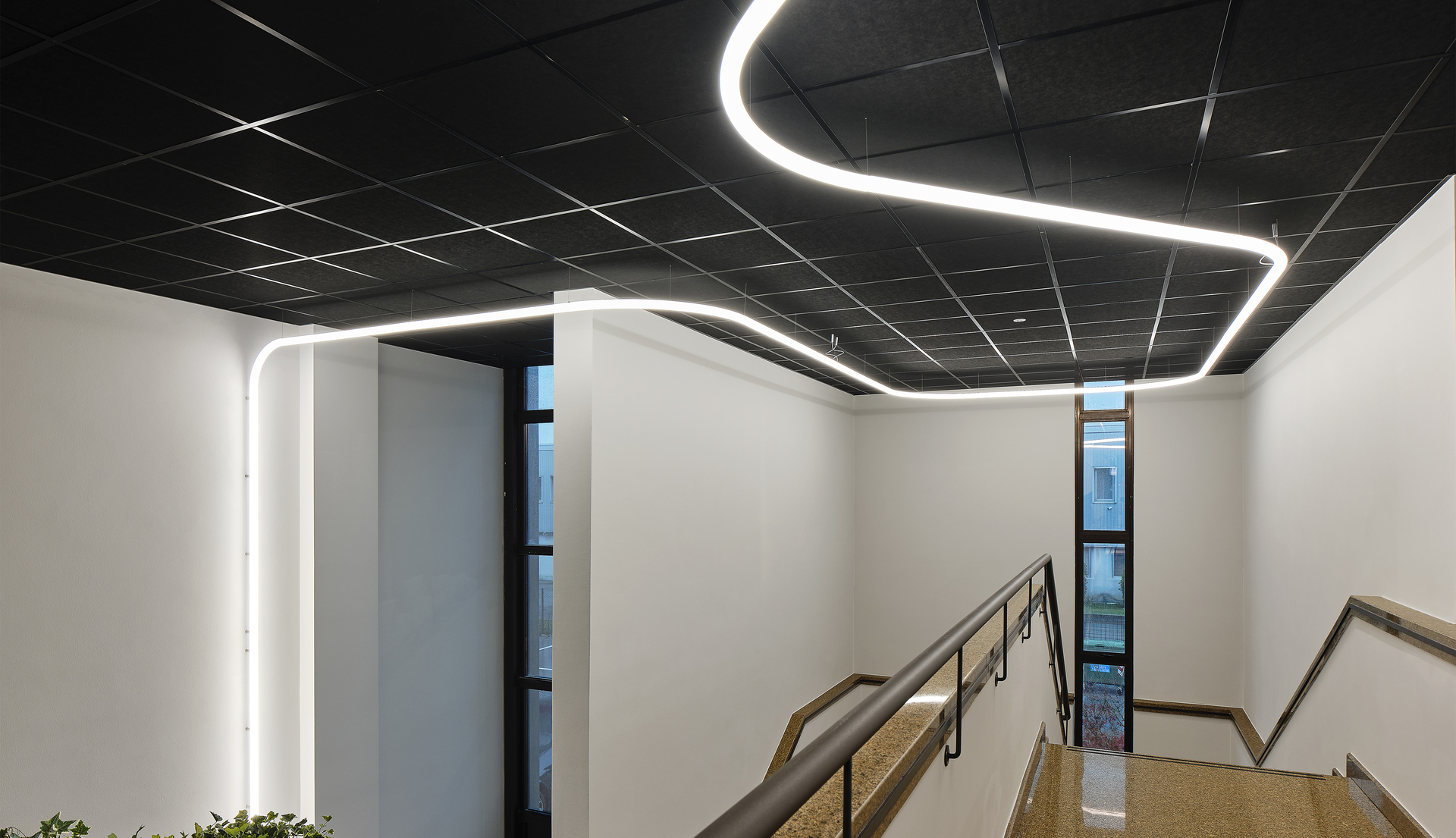 Curved continuous LED light strip installed on a dark grid ceiling, illuminating a modern stairwell with white walls.