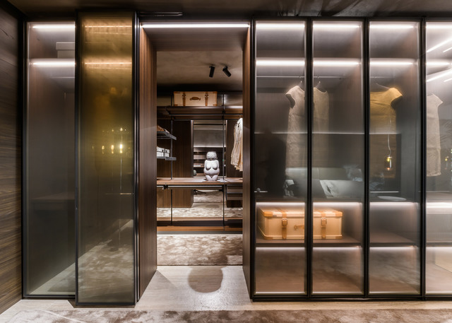 Luxury walk-in closet with glass sliding doors, dark wood shelving, and warm LED strip lighting illuminating display cases and interior.