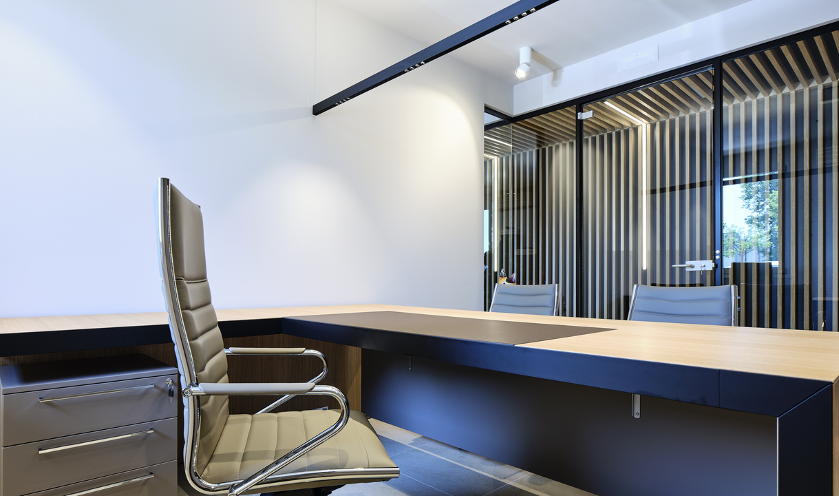 Modern office with L-shaped wood desk, executive chairs, track lighting on ceiling, and glass partition wall with vertical slats.