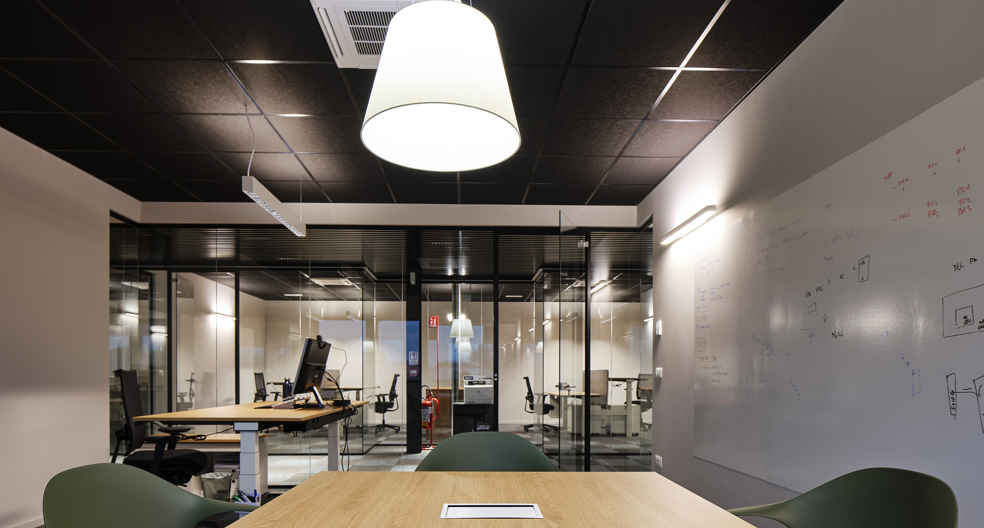 Modern office with large white conical pendant lamp over a wooden table, glass partitions, and workstations in background.