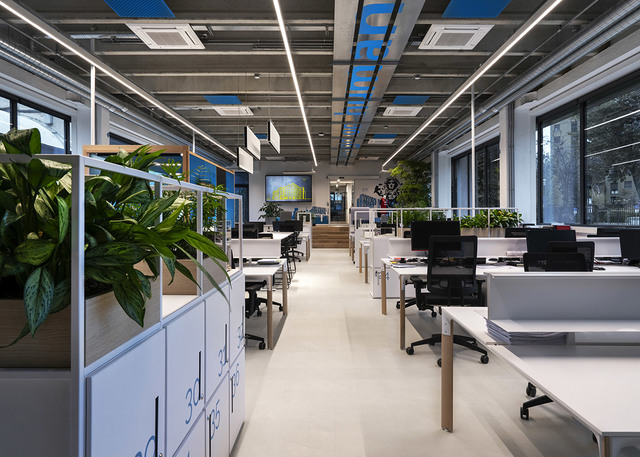 Ufficio open space moderno con lampade lineari LED Artemide a soffitto che illuminano postazioni di lavoro, scrivanie bianche, sedie ergonomiche e piante verdi.
