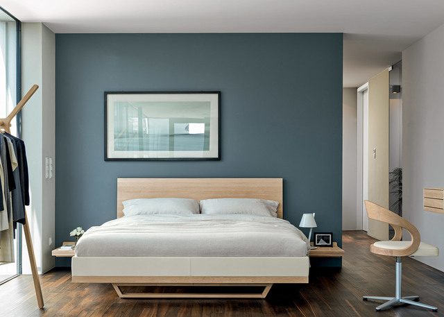 Camera da letto moderna con parete blu-verde, testiera in legno chiaro, biancheria bianca, piccole lampade da comodino e sedia scultorea