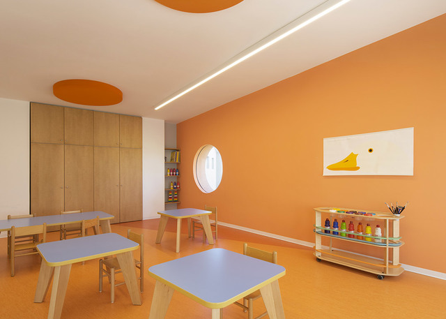 Aula per bambini luminosa con pareti arancioni, tavolini e sgabelli, illuminata da una luce a soffitto lineare LED a incasso.