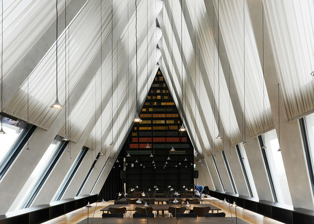 Interno di grande biblioteca con soffitto triangolare imponente, luci a sospensione sopra lunghi tavoli di lettura, scaffali colorati sullo sfondo