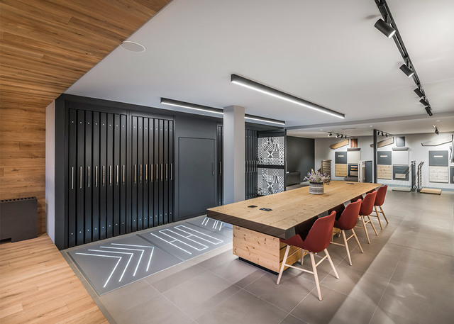Showroom moderno con luci lineari a soffitto, faretti su binario, tavolo riunioni in legno con sedie rosse e illuminazione decorativa a pavimento.