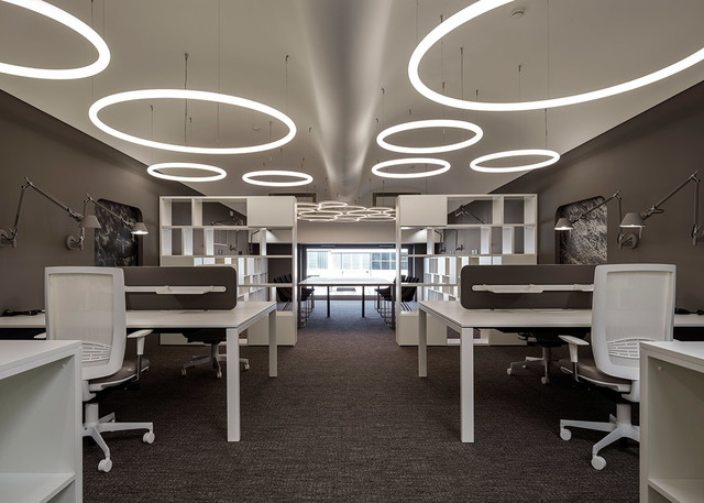 Ufficio moderno con file di scrivanie bianche e lampade a sospensione circolari Artemide disposte sul soffitto, creando un'illuminazione ambientale luminosa.
