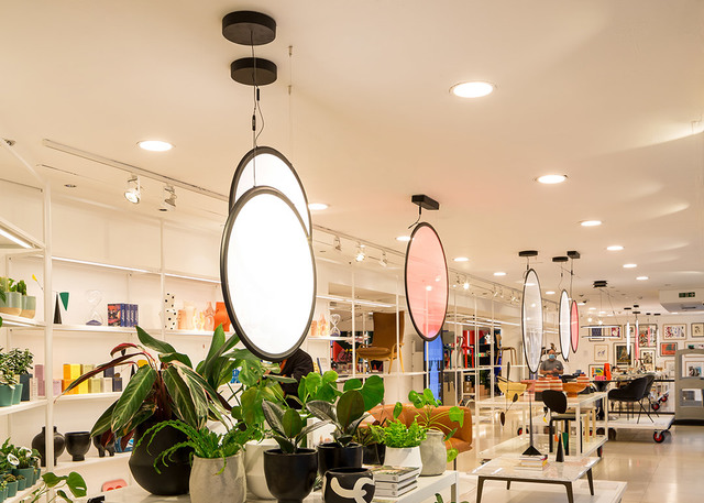 Interno di negozio con lampade a sospensione ovali Artemide in toni bianchi e rosa, sospese da strutture nere, sopra scaffali con piante e decorazioni.
