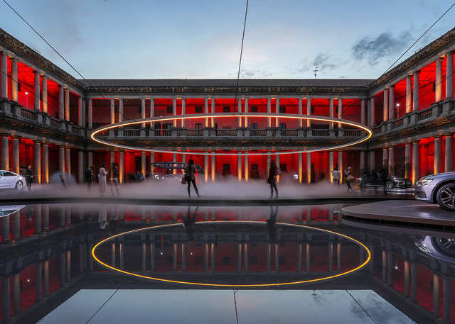 Grande anello luminoso circolare sospeso su un cortile nebbioso, riflesso in una vasca, con edificio neoclassico illuminato di rosso al crepuscolo