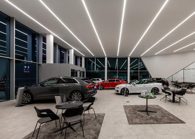 Showroom automobilistico moderno con luci lineari a LED a soffitto che illuminano veicoli di lusso su pavimento in cemento levigato.