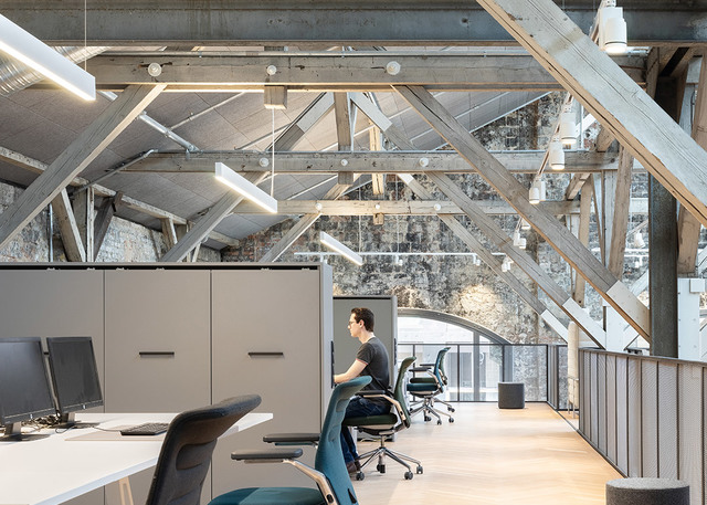 Spazio ufficio moderno con lampade lineari a sospensione Artemide sotto capriate in legno a vista, lavoratori alle scrivanie con sedie ergonomiche verde acqua