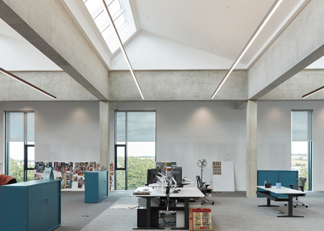 Ufficio open space moderno con soffitti alti in cemento, lucernari, luci lineari a LED a soffitto, postazioni con monitor e ampie finestre sul verde.