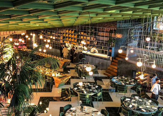 Elegant restaurant interior with globe pendant lights hanging over dark marble tables, green chairs, tropical plants, and a bar area.