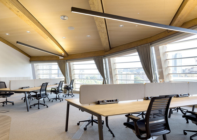 Modern office workspace with linear pendant LED light fixture suspended from wooden ceiling beams, illuminating open-plan desks and chairs.