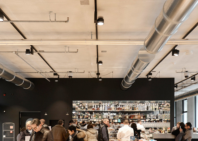 Interno di un caffè affollato con soffitto industriale a vista, condotti HVAC argentati e faretti Artemide neri su binario che illuminano un'area bar.