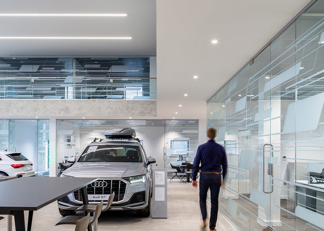 Showroom Audi con luci a soffitto incassate e lineari che illuminano un'area espositiva con SUV Q7, pareti in vetro e spazio di lavoro aperto.