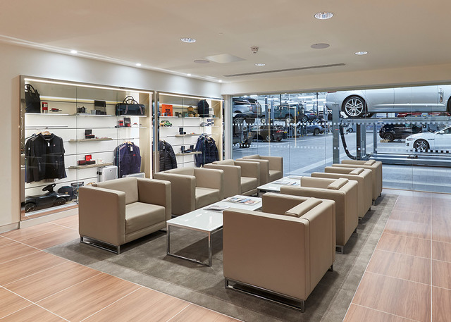 Luxury car dealership lounge with recessed ceiling lights, beige armchairs on a rug, display shelves with accessories, and showroom visible through glass.