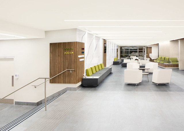 Atrio ospedaliero moderno con luci lineari a soffitto incassate, sedute bianche, pareti con pannelli in legno e cuscini verde-giallo.