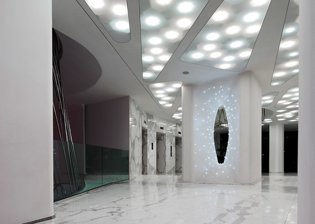 Ingresso moderno con luci LED circolari a incasso nel soffitto che illuminano un pavimento in marmo e un pannello decorativo ovale luminoso a parete.