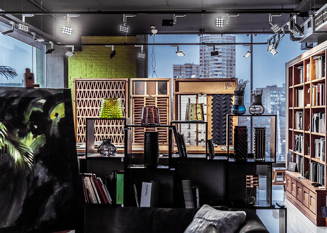 Modern interior showroom with track lighting on ceiling, wooden shelving units displaying decorative objects, bookcase, and city view.