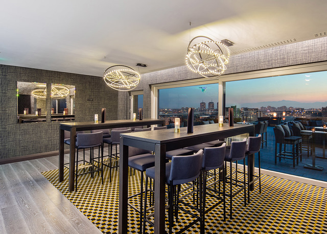 Bar sul tetto con due lampade a sospensione circolari LED Artemide che illuminano tavoli alti e sgabelli, con vista panoramica sulla città al tramonto.