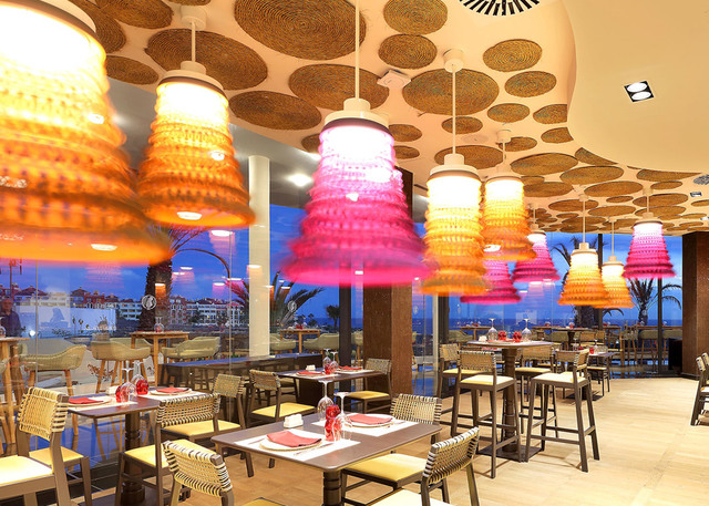 Restaurant interior with colorful tiered pendant lamps in orange, pink, and yellow hanging from a decorative ceiling with circular woven panels