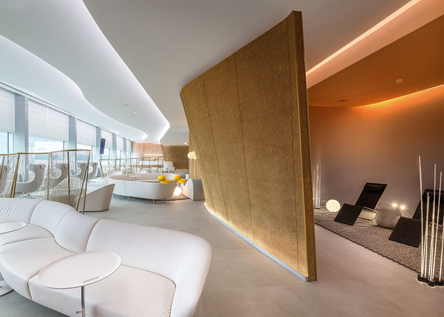 Lounge moderno con divani bianchi curvi, illuminazione indiretta calda a soffitto, parete divisoria dorata e lampade da terra in zona relax
