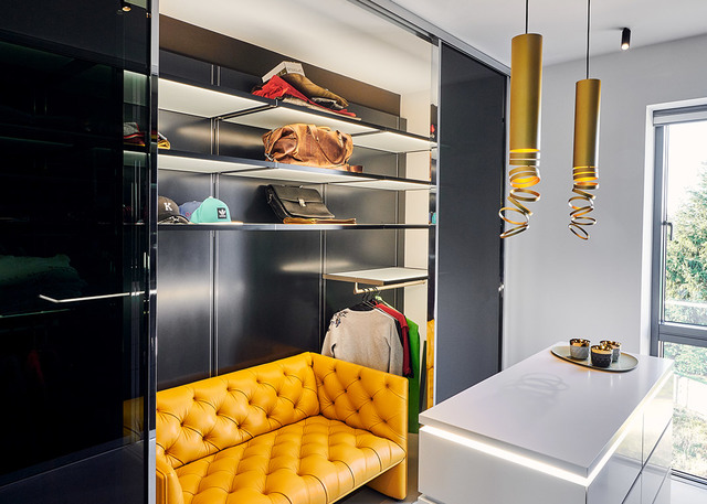 Cabina armadio boutique con lampade a sospensione a spirale dorata, divano trapuntato giallo, scaffalature scure con accessori e bancone bianco.